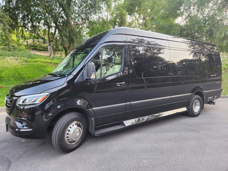 This black Mercedes Sprinter van, an RV rental, is parked on asphalt with green trees reflecting on its glossy finish.
