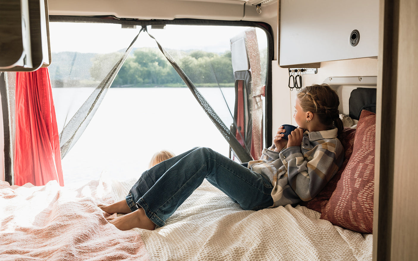 RV rental comfort: A girl in a plaid shirt sips from a mug, gazing at a lake view from the bed. Another child's head is nearby.