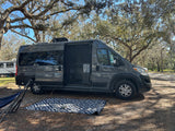 A sleek grey RV rental, with its side door open to an outdoor mat & hammock, is perfectly set in a serene, tree-filled campsite.