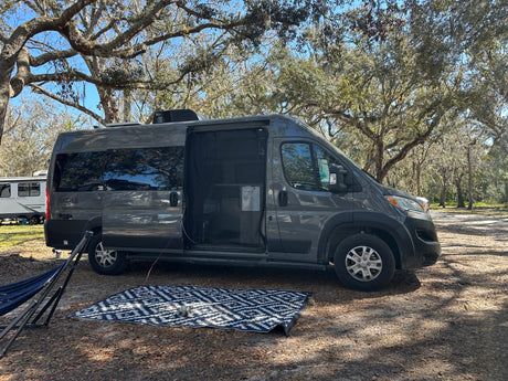 A sleek grey RV rental, with its side door open to an outdoor mat & hammock, is perfectly set in a serene, tree-filled campsite.