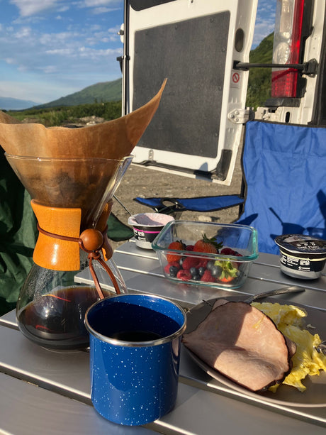 RV rental morning: Freshly brewed coffee, scrambled eggs, ham, berries, & yogurt on a camping table with mountain backdrop.