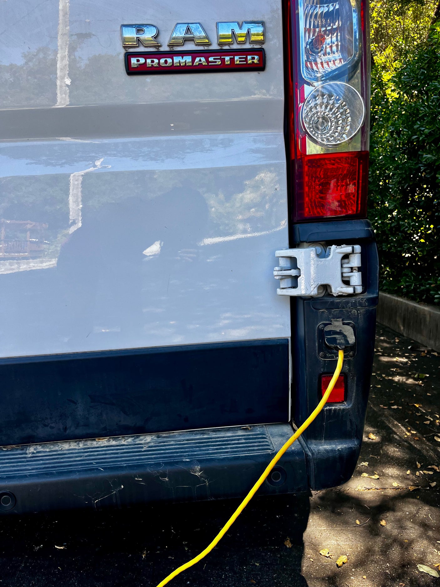 Rear view of an RV rental: a white Ram ProMaster van with a yellow power cord connected to its power inlet near the taillight.