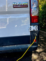 Rear view of an RV rental: a white Ram ProMaster van with a yellow power cord connected to its power inlet near the taillight.