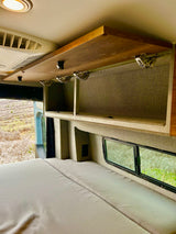 Interior of an RV rental: a comfortable bed, open overhead storage cabinet, and a window framing a natural landscape.