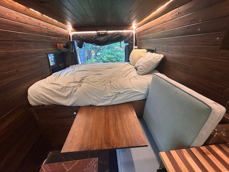 Cozy RV rental interior: A wood-paneled camper van with a comfy bed, table, and scenic window view. — photo 18