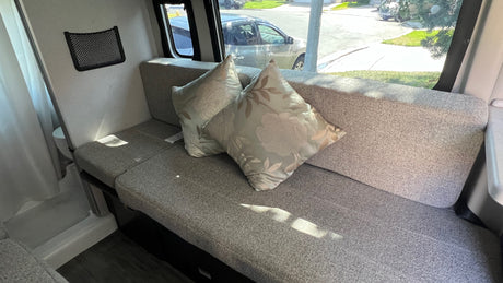 Cozy RV rental interior: grey sofa with floral pillows, window views, and a small storage net.