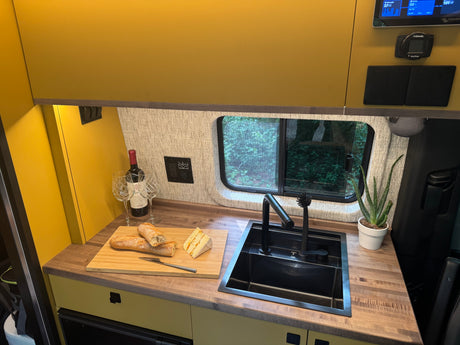 An RV rental's cozy kitchenette features a black sink, wood counter, control panel, and delicious snacks for travel.