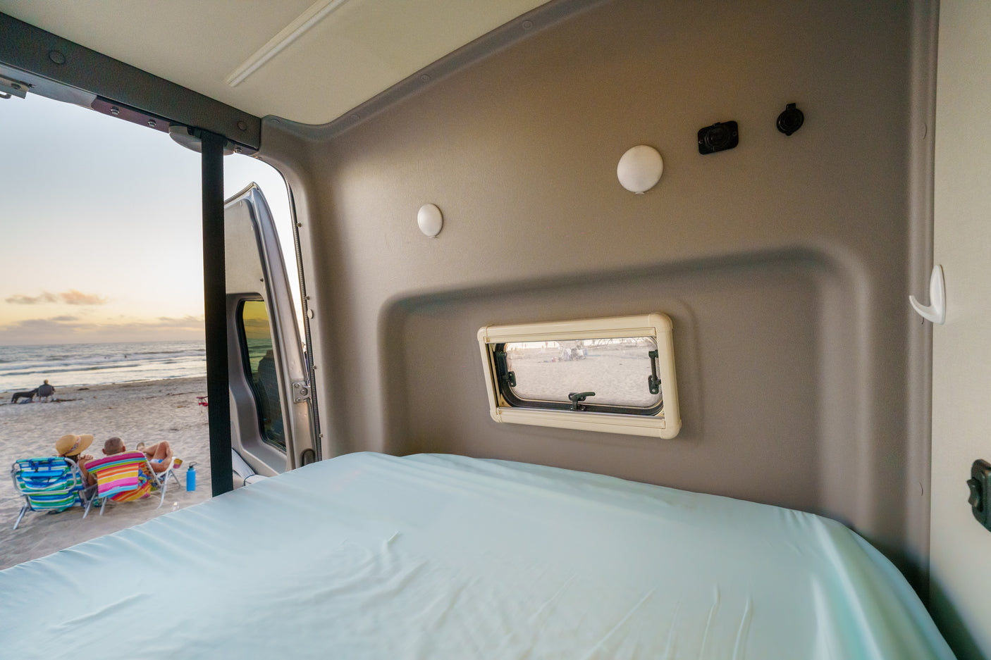 Relax in this RV rental! Comfy bed faces open rear door, revealing a beach sunset with people.