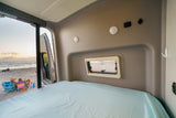 Relax in this RV rental! Comfy bed faces open rear door, revealing a beach sunset with people.