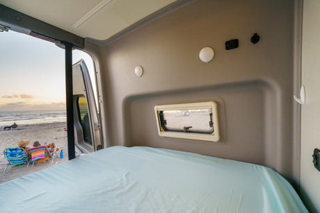 Relax in this RV rental! Comfy bed faces open rear door, revealing a beach sunset with people.