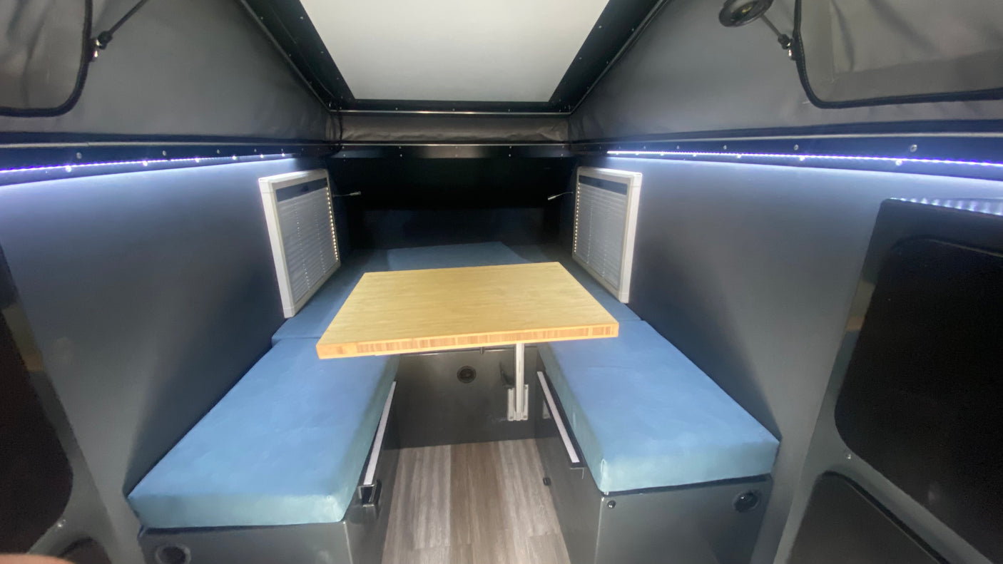 Inside this **RV rental**: a bright, compact dining space with blue benches, a wooden table, and perimeter LED lighting. Ideal for travel.