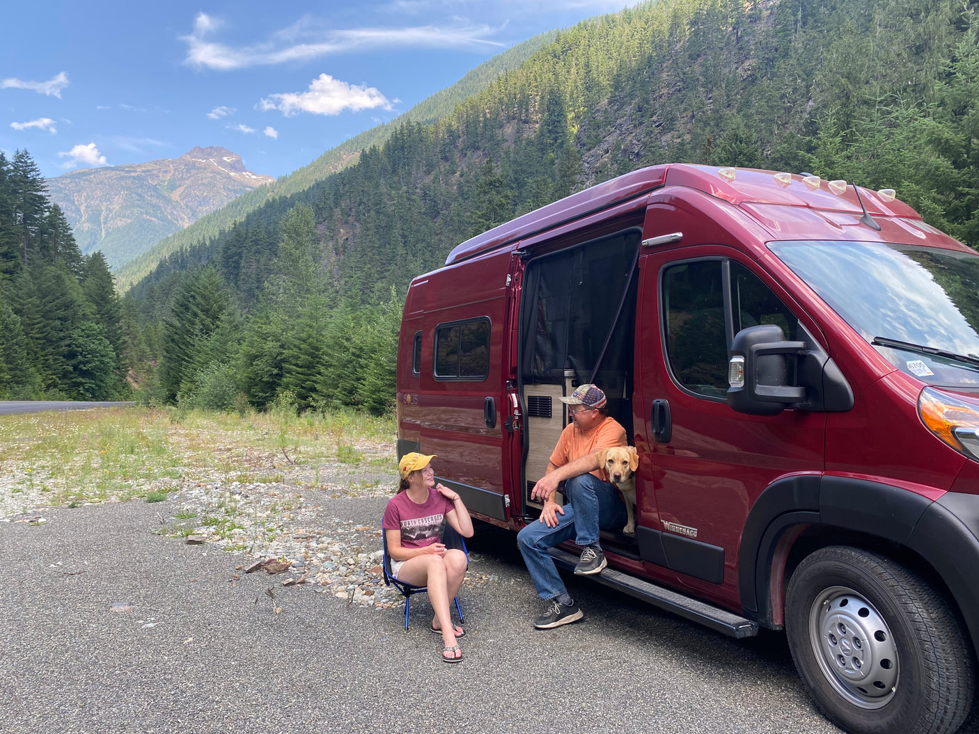 An RV rental trip! A couple and their dog relax by a red camper van in a beautiful mountain forest setting.