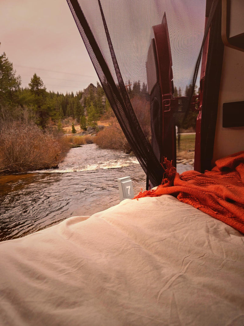  perfect for outdoor travel.|Riverside RV rental: A red camper van with a camp chair and fire pit by a flowing river at dusk.|Cozy RV rental bed with an orange blanket and bug net — photo 3
