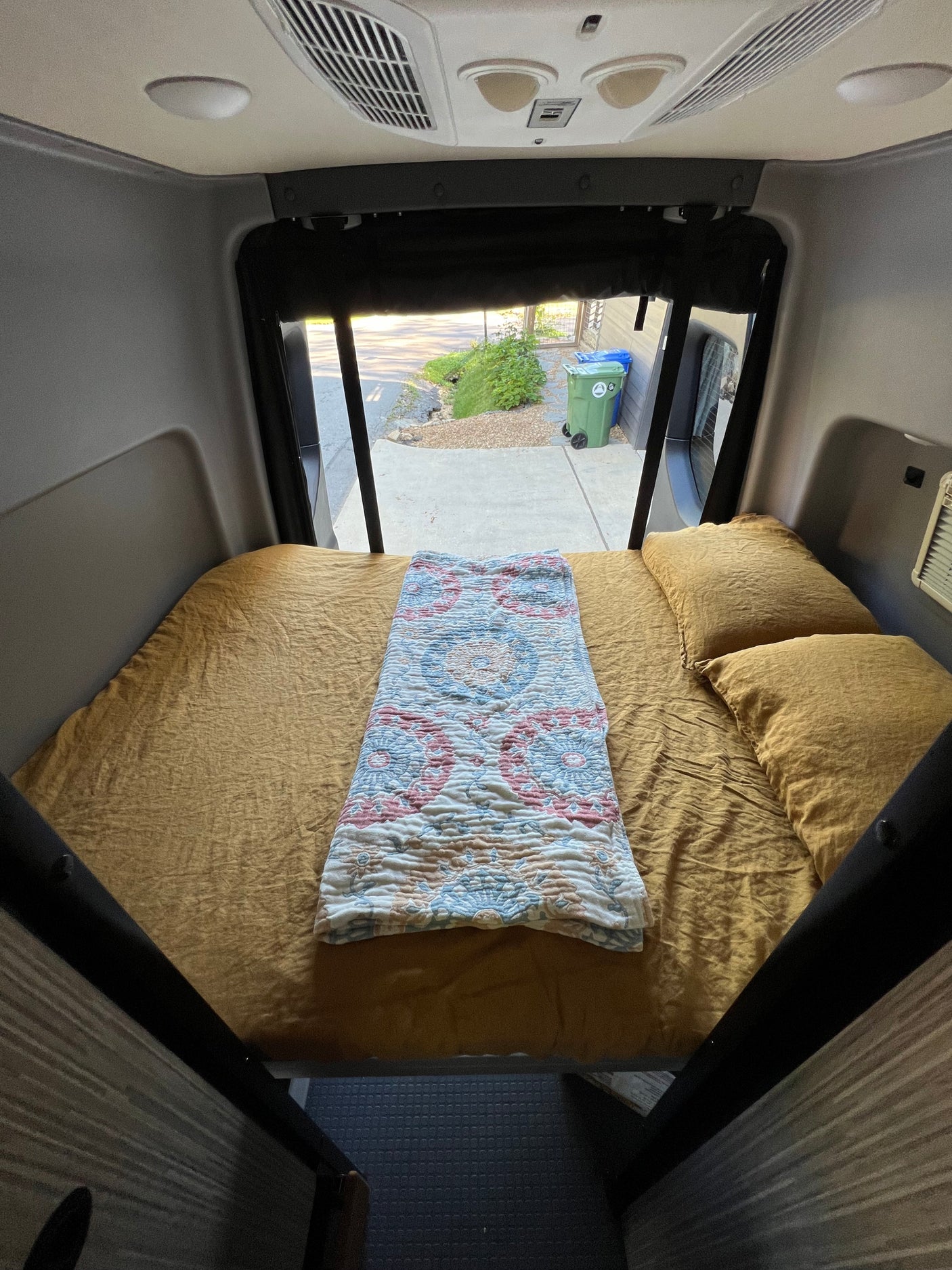 Cozy RV rental bed with mustard yellow sheets & patterned blanket inside a camper van. View out the back window.