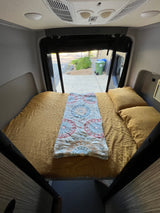 Cozy RV rental bed with mustard yellow sheets & patterned blanket inside a camper van. View out the back window.