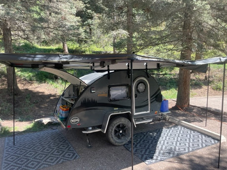This compact teardrop RV rental, featuring a full awning and outdoor mat, is perfectly set up in a lush, wooded campsite.