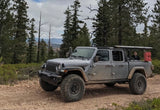 A rugged gray Jeep Gladiator pickup truck on a dirt trail in a forest, with a person in the back. Like an rv rental, perfect for adventure!