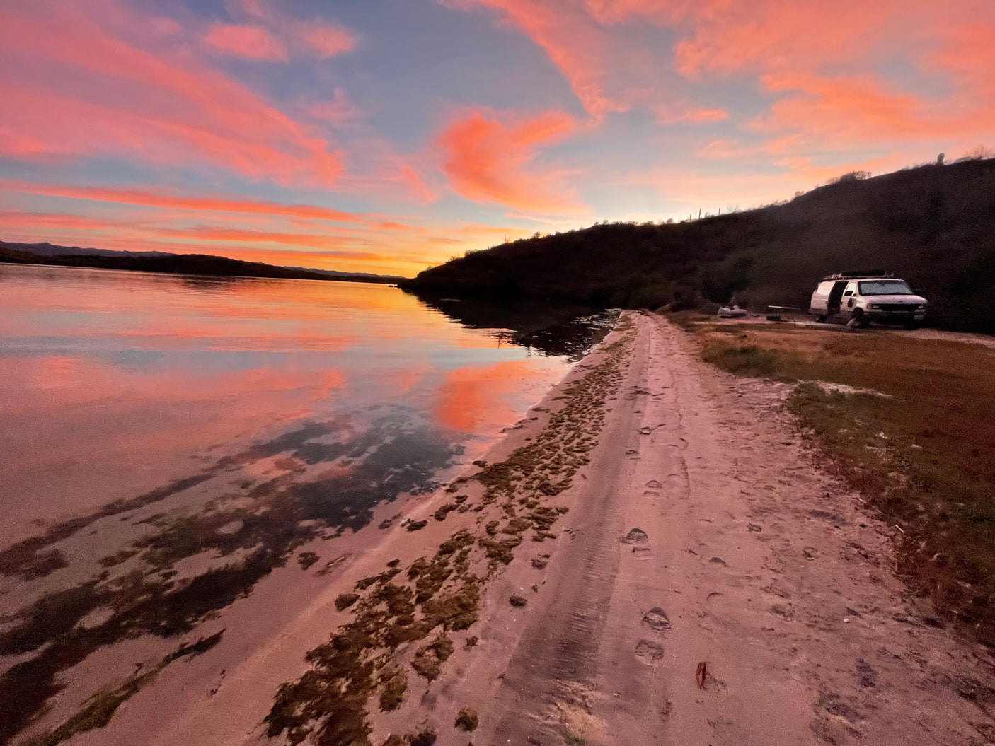 A breathtaking sunset with pink and orange clouds reflected on calm water. An RV rental van is parked on a sandy path beside a dark hill, inviting travel.