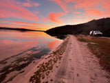 A breathtaking sunset with pink and orange clouds reflected on calm water. An RV rental van is parked on a sandy path beside a dark hill, inviting travel.
