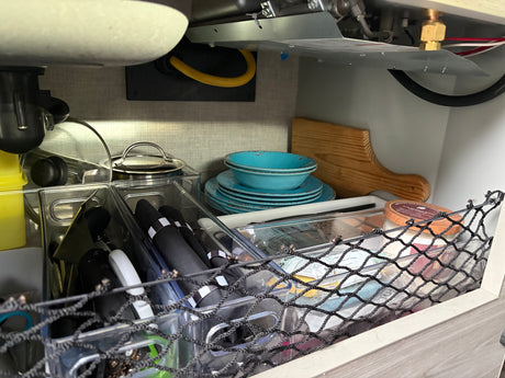 Compact kitchen storage under the sink in an RV rental, with stacked blue dishes, utensils, and organizers.