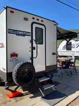 A white Yellowstone travel trailer, perfect for an rv rental, is parked at a sunny campsite with its awning out and outdoor seating.