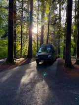 RV rental camper van parked in a sun-dappled forest campsite, surrounded by tall trees. Perfect nature escape.
