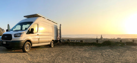 Enjoying beach camping in a white Ford Transit RV rental, parked on sand with solar panels, overlooking the ocean at sunset.
