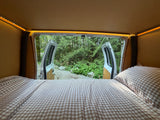 Cozy RV rental bed with an open view to a lush forest from inside. Experience comfortable camping with nature at your doorstep.