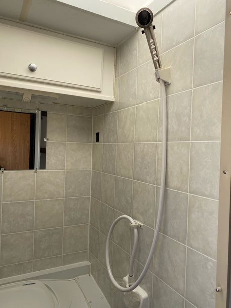 Clean, compact RV rental shower with neutral tiled walls, a handheld showerhead, and upper storage cabinet.
