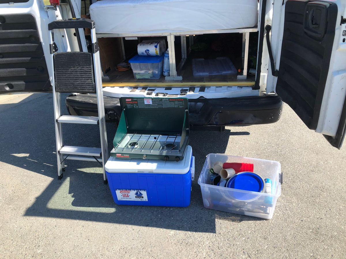 Rear view of an RV rental showing interior storage, a ladder, Coleman camping stove on a cooler, and a bin of kitchen supplies. — photo 12