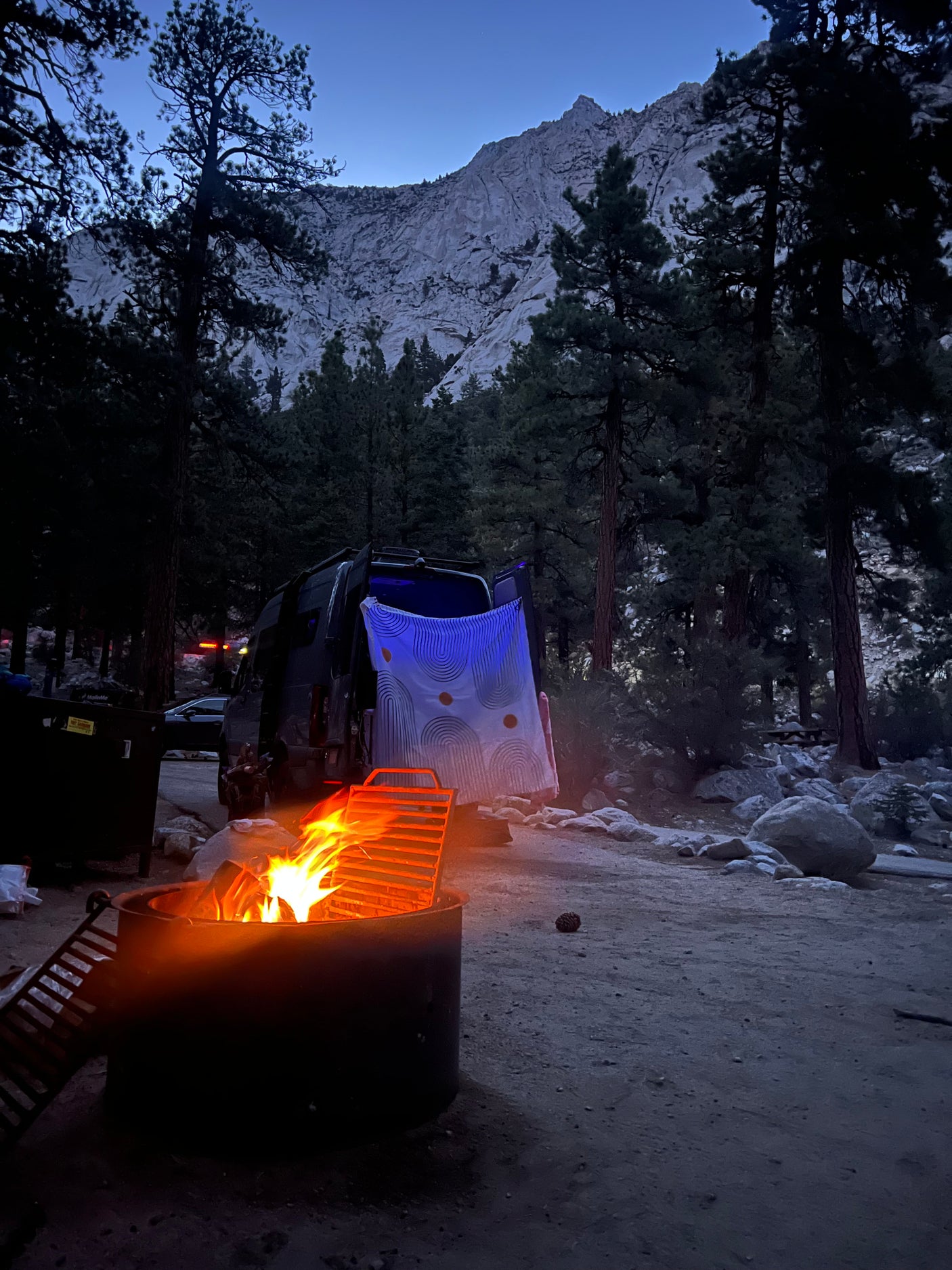 Campsite at dusk with an RV rental, warm campfire, tall pine trees, and a grand mountain.