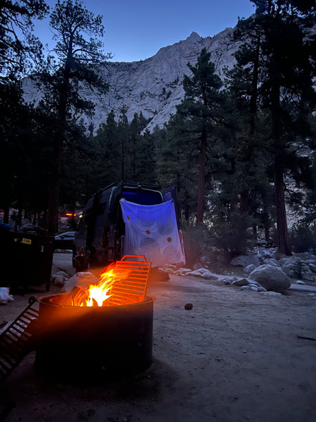 Campsite at dusk with an RV rental, warm campfire, tall pine trees, and a grand mountain.