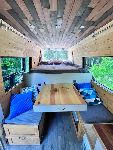 Spacious rv rental interior: wooden walls/ceiling, comfy bed, dining table, and windows with forest views.