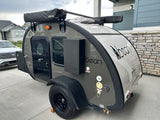 Gray and white Bean teardrop trailer, a compact RV rental, with awning and roof fan, parked on a driveway.