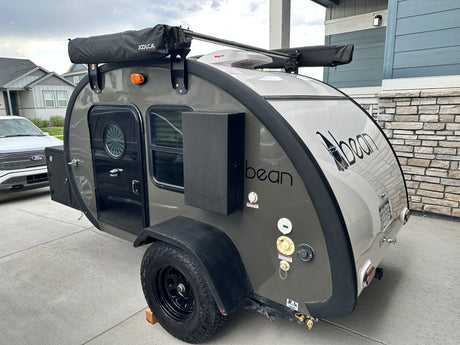 Gray and white Bean teardrop trailer, a compact RV rental, with awning and roof fan, parked on a driveway.