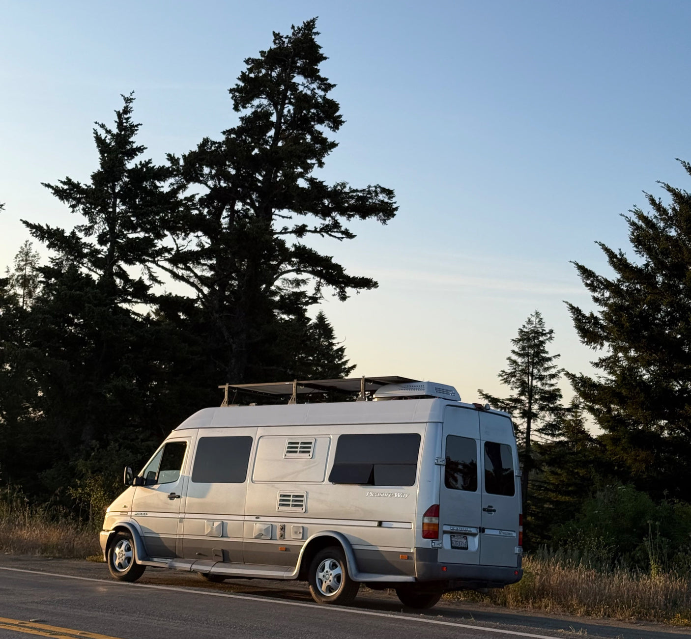 Silver Pleasure-Way Sprinter RV rental parked roadside by tall trees under a clear sky, ready for adventures.