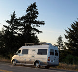 Silver Pleasure-Way Sprinter RV rental parked roadside by tall trees under a clear sky, ready for adventures.