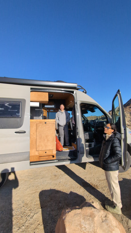 This RV rental camper van offers adventure! Two people stand by a gray van with wooden interior, under a clear blue sky.