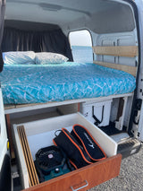 Cozy camper van bed with ocean view. Features under-bed storage, cooler, and fan for your RV rental adventure.
