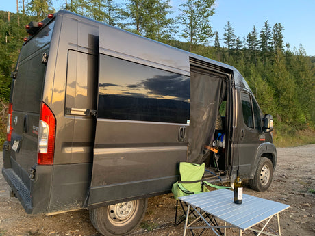 Relaxing outside a gray RV rental camper van, set up with a table, wine, and chair amidst a beautiful forest backdrop.