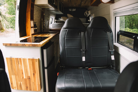 RV rental's cozy interior: kitchen with induction cooktop, sink, fridge, and two passenger seats with seatbelts.