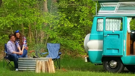 Relaxing RV rental getaway: Couple by a campfire, vintage teal camper van in green nature.