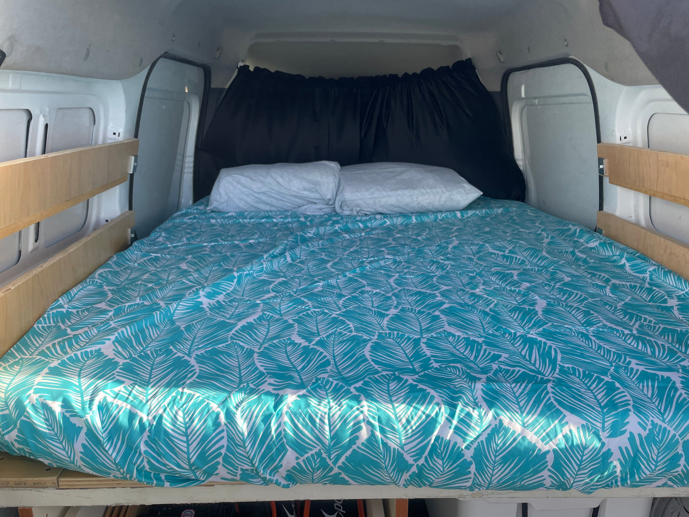 Inside an RV rental, a cozy campervan bed is made up with vibrant teal, leaf-patterned bedding and privacy curtains.