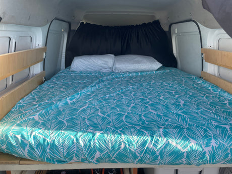Inside an RV rental, a cozy campervan bed is made up with vibrant teal, leaf-patterned bedding and privacy curtains.
