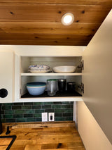 Compact RV rental kitchen with a wood ceiling, open cabinet showing dishes, green tile backsplash, & wooden counter.