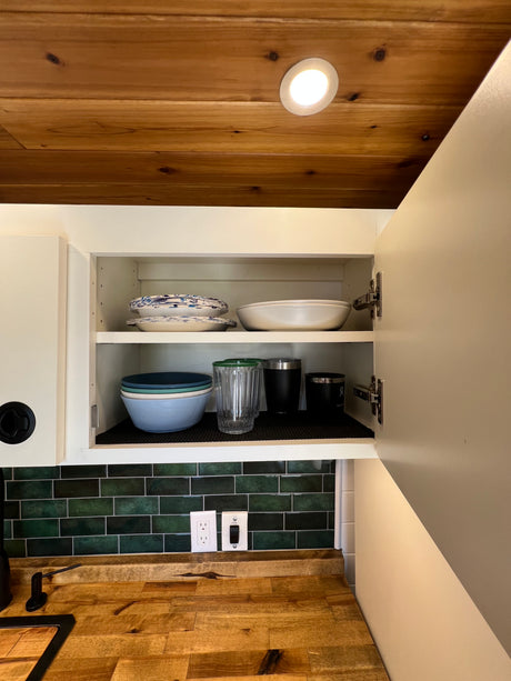 Compact RV rental kitchen with a wood ceiling, open cabinet showing dishes, green tile backsplash, & wooden counter.