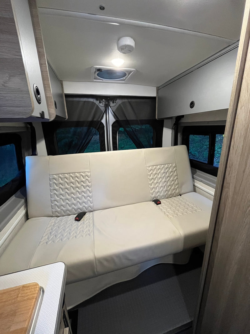 Inside this modern rv rental: a light-colored bench seat, overhead cabinets, and rear windows. Perfect camper van living space. — photo 4