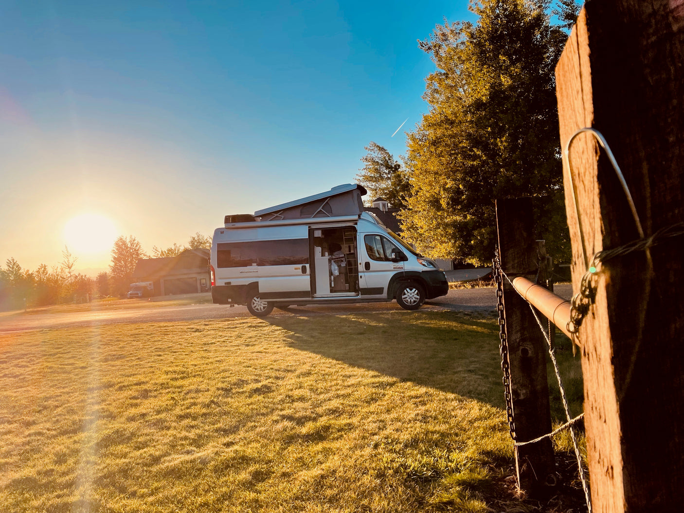 Experience sunrise with an RV rental! A camper van parked in a sunny field with open door, pop-top, and a wooden fence post.