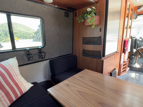 Inside a comfy RV rental: dining table, window with scenic mountain road, seating area, and a dog visible in the front.