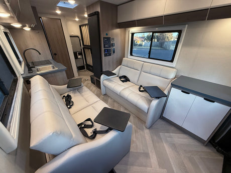 Modern RV rental interior: bright, spacious living area with two white recliners, kitchen, and wood-look flooring.